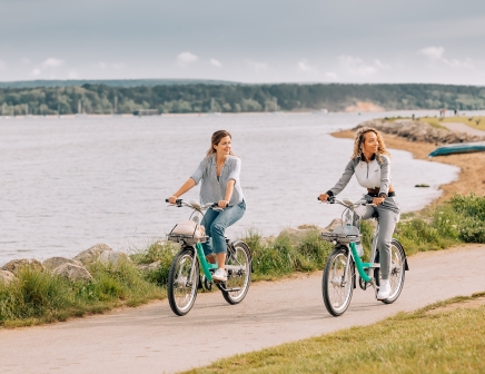 Two female riders using Beryl bikes along a coastal path. Both riders are looking to their left at something off camera.
